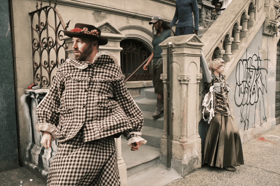 Un hombre vestido con un traje de cuadros y un sombrero decorado camina por la acera, mientras otras personas están en las escaleras de un edificio con grafitis en las paredes.